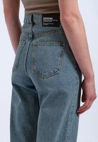 Light blue denim jeans with a high waist, five pockets, and yellow stitching. Features a black label with logo on the back.