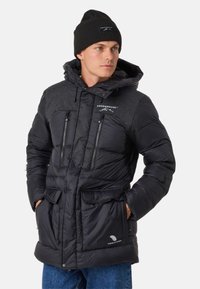 Black puffer jacket with a quilted upper section, two zippered chest pockets, and large front pockets; paired with a black knit beanie.