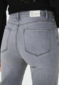 Šedé denimové džíny s vysokým pasem, viditelnými zadními kapsami a štítkem s nápisem "modré denim." Jemná textura, klasický střih.