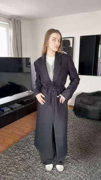 Μακρύ μαύρο παλτό με ζώνη που δένει, πλαϊνές τσέπες και κλείσιμο με κουμπιά· κατασκευασμένο από λείο ύφασμα, με κομψό, ραμμένο σχέδιο.