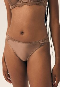 Biustonosz i koronkowy zestaw bielizny w jasnobrązowym kolorze, charakteryzujący się przezroczystą, gładką teksturą oraz misternymi koronkowymi detalami wzdłuż linii talii.