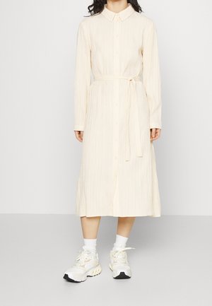 Roomkleurige, geribbelde shirtjurk met lange mouwen, knoopsluiting, ceintuur in de taille en een zoom tot op de knie. Draagt witte sneakers.