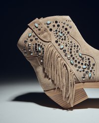 Botín de ante beige hasta el tobillo con flecos, adornado con tachuelas negra y turquesa, con un tacón de madera texturizada y un patrón decorativo.