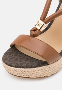 Gros plan sur une sandale compensée en cuir marron avec semelle en corde tissée, semelle intérieure à motifs et détail du logo doré MK sur la bride de cheville nouée.