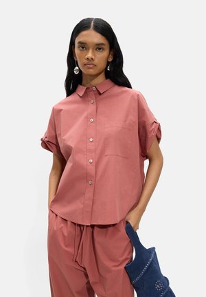 Kobieta ubrana w luźną, pudrowo-różową koszulę na guziki i pasujące spodnie, trzymająca małą niebieską torebkę z srebrnymi ćwiekami.