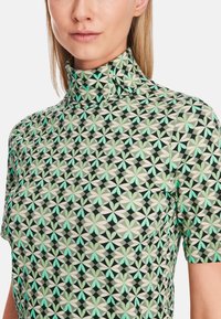 Kragenoberteil mit kurzen Ärmeln in einem geometrischen Blumenmuster in Grün, Schwarz und Creme. Verfügt über einen strukturierten Stoff mit taillierter Silhouette.