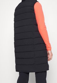 Svart vadderad väst med hög krage, med horisontella quiltade sektioner och en strömlinjeformad design. Bärs över en ljus korallfärgad långärmad topp.