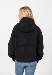 Hoodie noir court en tissu pelucheux, avec une grande capuche et des poignets côtelés. Le dos présente une finition texturée et douce.