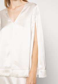 Femme portant un chemisier blanc en satin à col en V, avec un ourlet à franges et des manches fendues laissant apparaître le bras nu, sur un fond uni.