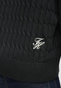 Černý pletený svetr s texturovaným vzorem copánku, žebrovanými manžetami a krémovým vyšívaným logem na přední straně.