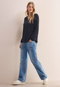 Mörk marinblå långärmad blus med v-ringning, tillsammans med ljusblå avslappnade jeans och vita sneakers, visad mot en neutral bakgrund.