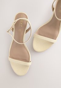 Sandales de couleur crème avec une tige en maille texturée, un design plat et une fine bride à la cheville. Semelle intérieure beige douce et détails de quincaillerie minimalistes.