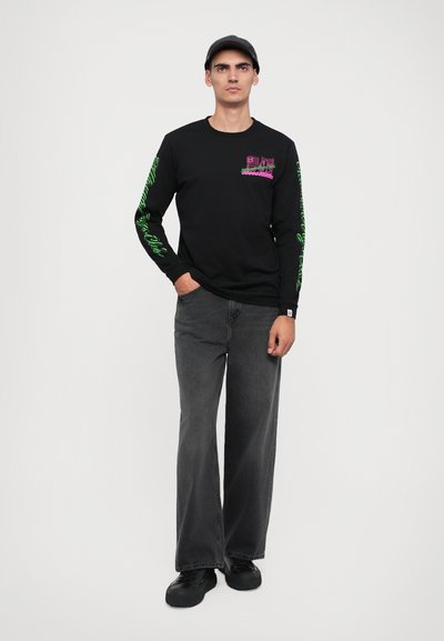 Camisola preta de manga longa com detalhes gráficos em rosa e verde, combinada com jeans cinza de corte largo e ténis pretos, contra um fundo simples.