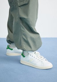 Person, der olivgrüne Hosen und weiße Adidas Stan Smith Sneakers mit grünem Fersenbereich auf einem hellblauen Teppich trägt.