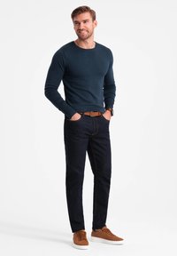 Marinblå långärmad skjorta, mörka jeans med gula sömmar, bruna mockaskor. Modellen står med händerna i fickorna mot en vit bakgrund.
