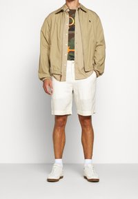 Beżowa, lekka kurtka, koszulka w kamuflaż, białe szorty i białe sneakersy z jasnobrązowymi podeszwami. Casualowy, luźny krój z subtelnym brandingiem.