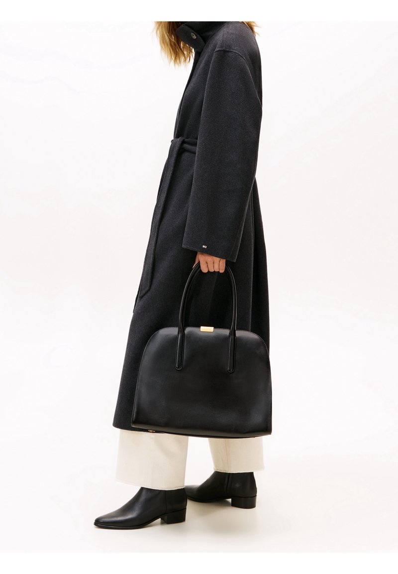 Femme en manteau long foncé et pantalon clair tenant un grand sac à main en cuir noir avec deux poignées, portant des bottines noires.