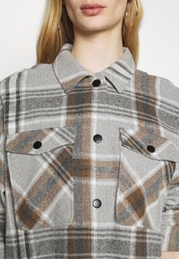 Gray plaid button-up shirt made of soft fabric, featuring brown and white accents, a collar, and two chest pockets with black buttons.
