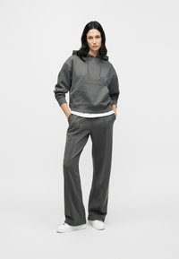 Γκρι φούτερ με κουκούλα, εμπρόσθια τσέπη και ρυθμιζόμενα κορδόνια, συνδυασμένο με γκρι παντελόνι φαρδιάς γραμμής που ταιριάζει. Φορεμένο με λευκά αθλητικά παπούτσια.