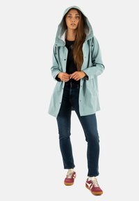 Chaqueta impermeable de color azul claro con capucha, cierre frontal a presión y forro interior gris en contraste, combinada con jeans oscuros y zapatillas rojas.