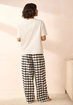 Frau steht barfuß drinnen, trägt ein schlichtes weißes kurzärmeliges Shirt und schwarz-weiße karierte Weitbein-Hose, schaut weg.