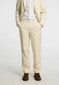 Beiga avslappnade byxor med elastisk midja och sidofickor, i kombination med en lätt, texturerad skjorta med knappar och mörka skor.