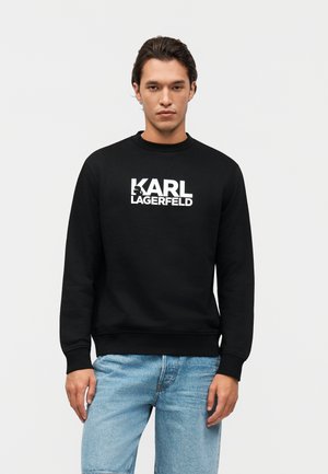 Uomo in piedi che indossa una felpa nera con la scritta "KARL LAGERFELD" in bianco e jeans azzurri chiari, su uno sfondo bianco uniforme.