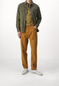 Camisa abotonada verde oliva, camiseta mostaza y pantalones de pana naranja, complementados con zapatillas blancas de acabado texturizado.