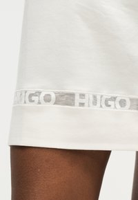 Άκρη λευκού ενδύματος με διαφανή λωρίδα από δίχτυ που φέρει το κείμενο "HUGO", φορεμένη σε μηρό με σκούρο δέρμα, κοντινή όψη.