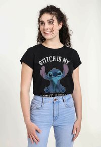 T-shirt noir à manches courtes arborant un personnage de dessin animé aux grandes oreilles et aux tons bleus, accompagné d'un texte gris disant "STITCH EST MON ANIMAL TOTEM."