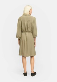 Khaki kleit, millel on kinnitatud vöökoht, pikad puhvseelikud ja veidi laienev alläär. Sile kangas, millel on peen sära. Mustad lamejalatsid.