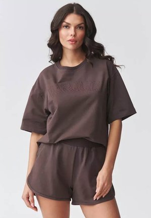 Kobieta o długich ciemnych włosach, ubrana w luźny ciemnobrązowy T-shirt i pasujące szorty, stojąca na jednolitym jasnym tle.