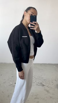 Giacca bomber nera con zip e vestibilità oversize, con il testo bianco "PECADOR" sul lato sinistro del petto, abbinata a pantaloni bianchi a vita alta.