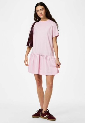 Mlada ženska z dolgimi temnimi lasmi, oblečena v ohlapno roza kratko rokavico, bordo superge in usklajeno torbico čez ramo.