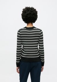 Πλεκτό πουλόβερ με ασπρόμαυρες ρίγες και ανάγλυφη υφή, με στρογγυλή λαιμόκοψη και μακριά μανίκια, συνδυασμένο με παντελόνι σκούρου χρώματος.