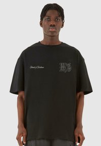 Tricou oversize din bumbac negru, cu mâneci scurte, guler rotund și grafica subtilă pe piept și mânecă. Textură netedă.