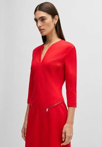 Rotes Kleid mit V-Ausschnitt, Dreiviertelärmeln und Reißverschlüssen an den Hüften. Glatte Textur und figurbetontes Design.