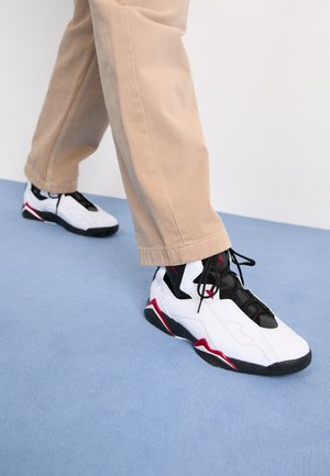 Person trägt beige Hose und weiße, schwarze und rote Sportschuhe und steht auf einem hellblauen Boden.