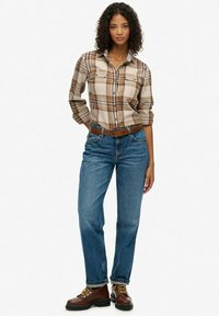 Chemise à carreaux marron et beige, jean en denim clair, ceinture marron, bottes marron avec lacets jaunes, coupe décontractée usée, tissu texturé.