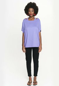 Luźna fioletowa bluzka z miękkiego materiału, z otwartą konstrukcją na ramionach. Połączona z czarnymi dopasowanymi spodniami i czarnymi sandałami.