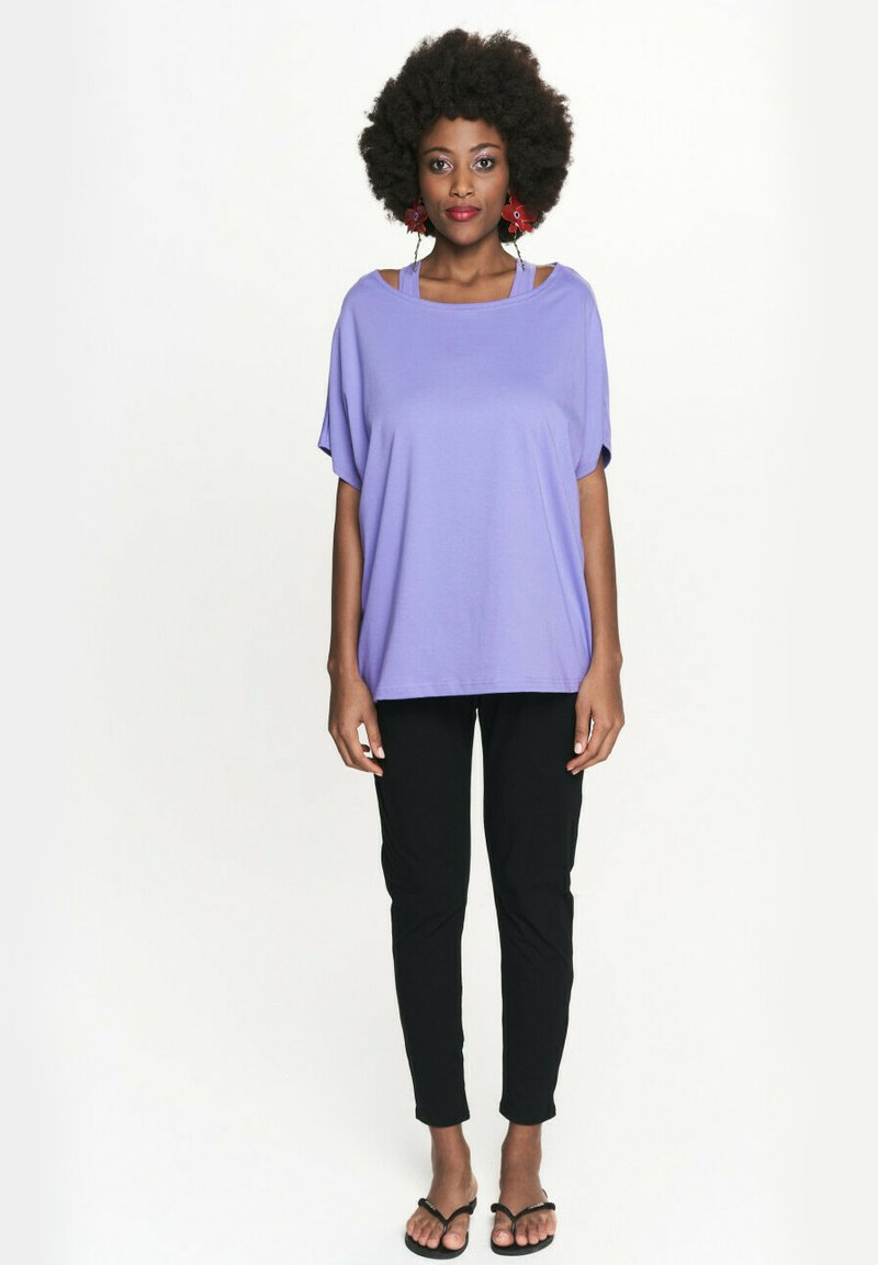 Luźna fioletowa bluzka z miękkiego materiału, z otwartą konstrukcją na ramionach. Połączona z czarnymi dopasowanymi spodniami i czarnymi sandałami.