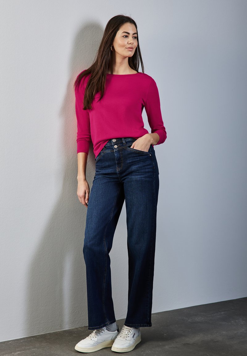 Fuchsia Langarmshirt kombiniert mit dunkelblauen weiten Jeans. Das Model trägt weiße Sneakers und zeigt einen lässigen und entspannten Stil.