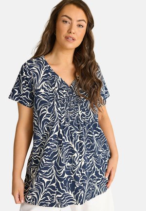 Kvinde iført en blå og hvid kortærmet bluse med abstrakt mønster, V-hals og knappet frontpanel.