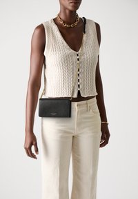 Saco a tiracolo em couro preto com um acabamento texturizado, alça de corrente dourada e um logótipo subtil. Usado com um top de malha em creme e calças claras.