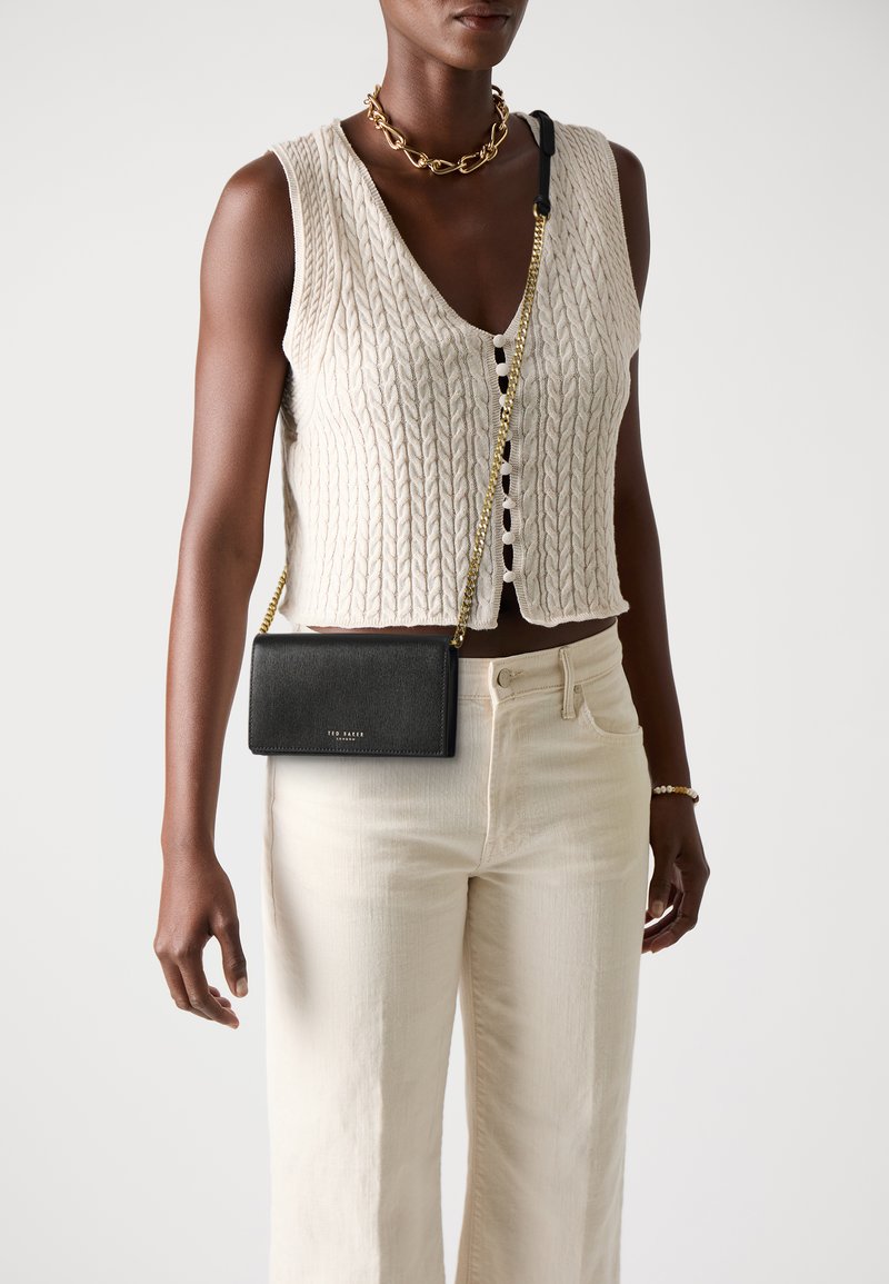 Saco a tiracolo em couro preto com um acabamento texturizado, alça de corrente dourada e um logótipo subtil. Usado com um top de malha em creme e calças claras.