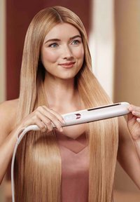 Witte stijltang met een roze accent, vastgehouden door een persoon met lang, recht blond haar. Glad oppervlak, ergonomisch ontwerp en snoer zichtbaar.