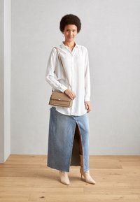Blusa bianca a maniche lunghe abbinata a una gonna maxi in denim blu chiaro con uno spacco davanti. Una borsa beige e stivaletti color crema completano il look.