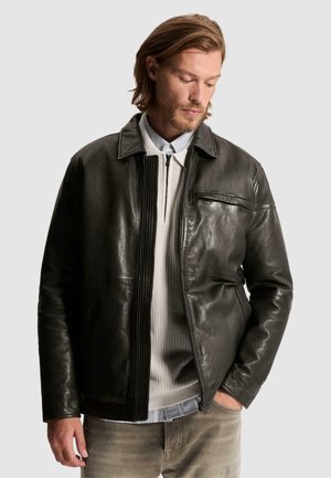 Homme aux cheveux mi-longs portant une veste en cuir noire par-dessus un pull blanc zippé et une chemise bleu clair, debout avec une main dans la poche.