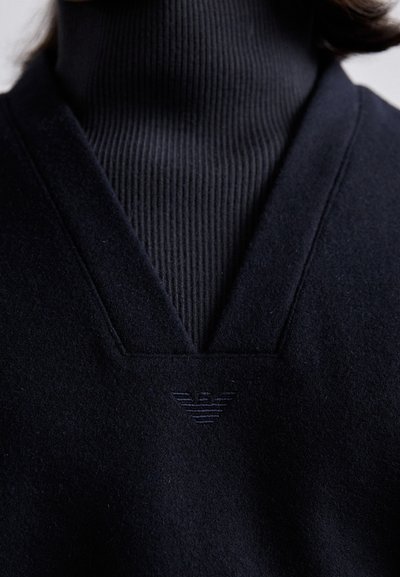Črna jopica z obliko V-izreza, pod katero je mehka rebrasta turtleneck. Na prsih je diskretno tonalno vezeno logo. Mehka tekstura.