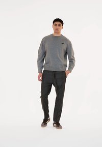 Sudadera gris con cuello redondo y pequeño logo; combinada con pantalones oscuros ajustados y zapatillas marrones con rayas blancas.